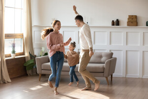 Cheery couple dancing with little daughter barefoot on wooden laminate floor with underfloor heating system in modern warm living room. New home, bank loan and lending, hobby and fun with kids concept